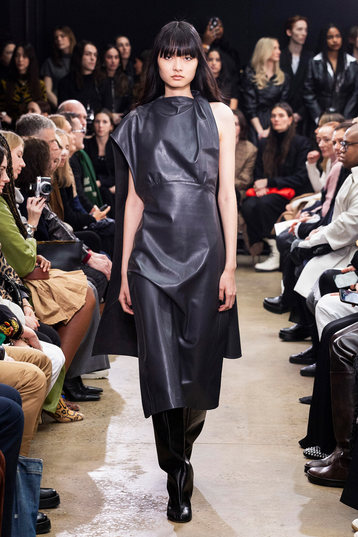 Runway model walks in a dark, sleeveless leather-like dress; audience seated on both sides watching.