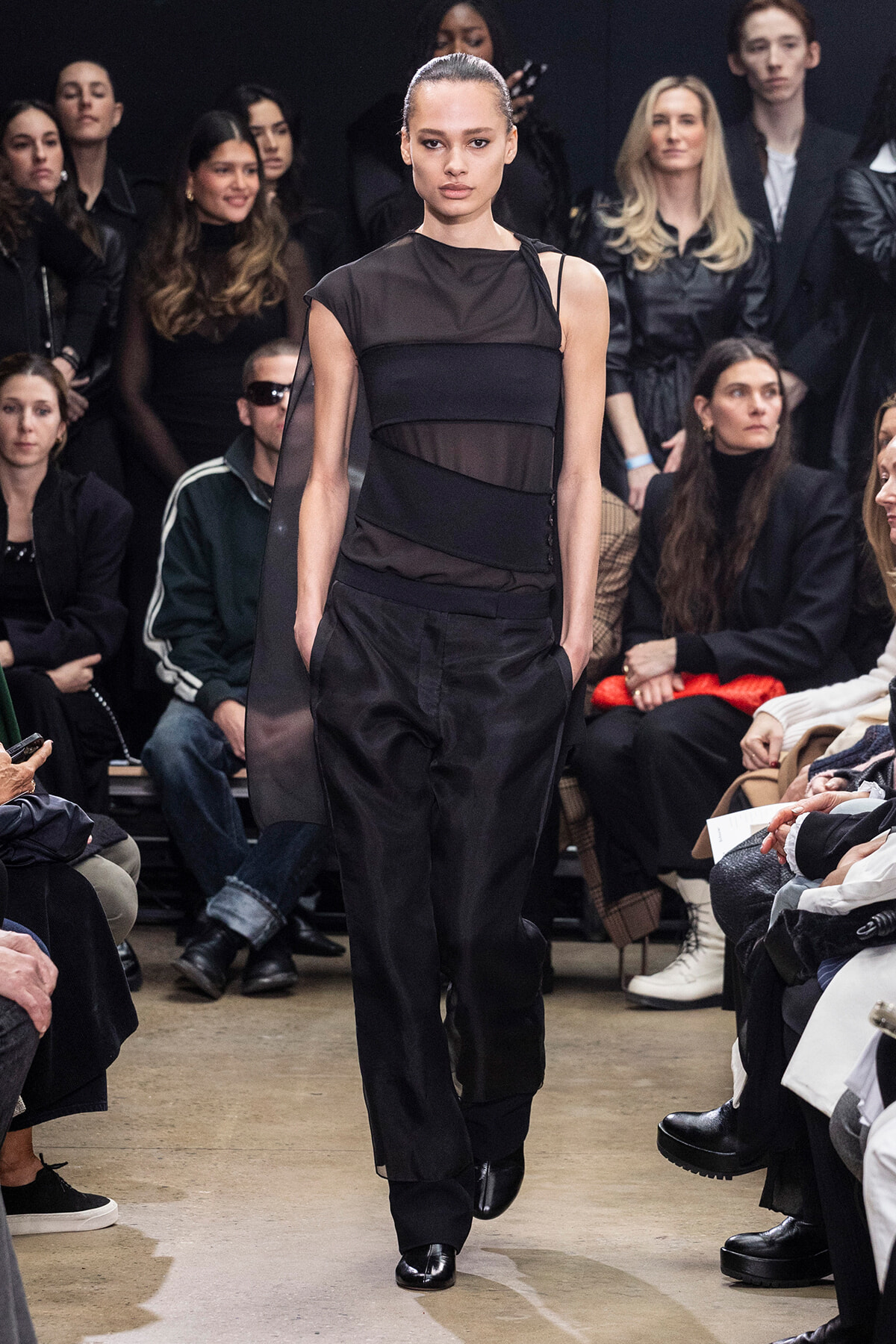 Model walking a fashion runway in a black, layered sheer and opaque outfit with hands in pockets; audience seated along the sides in dark clothing.