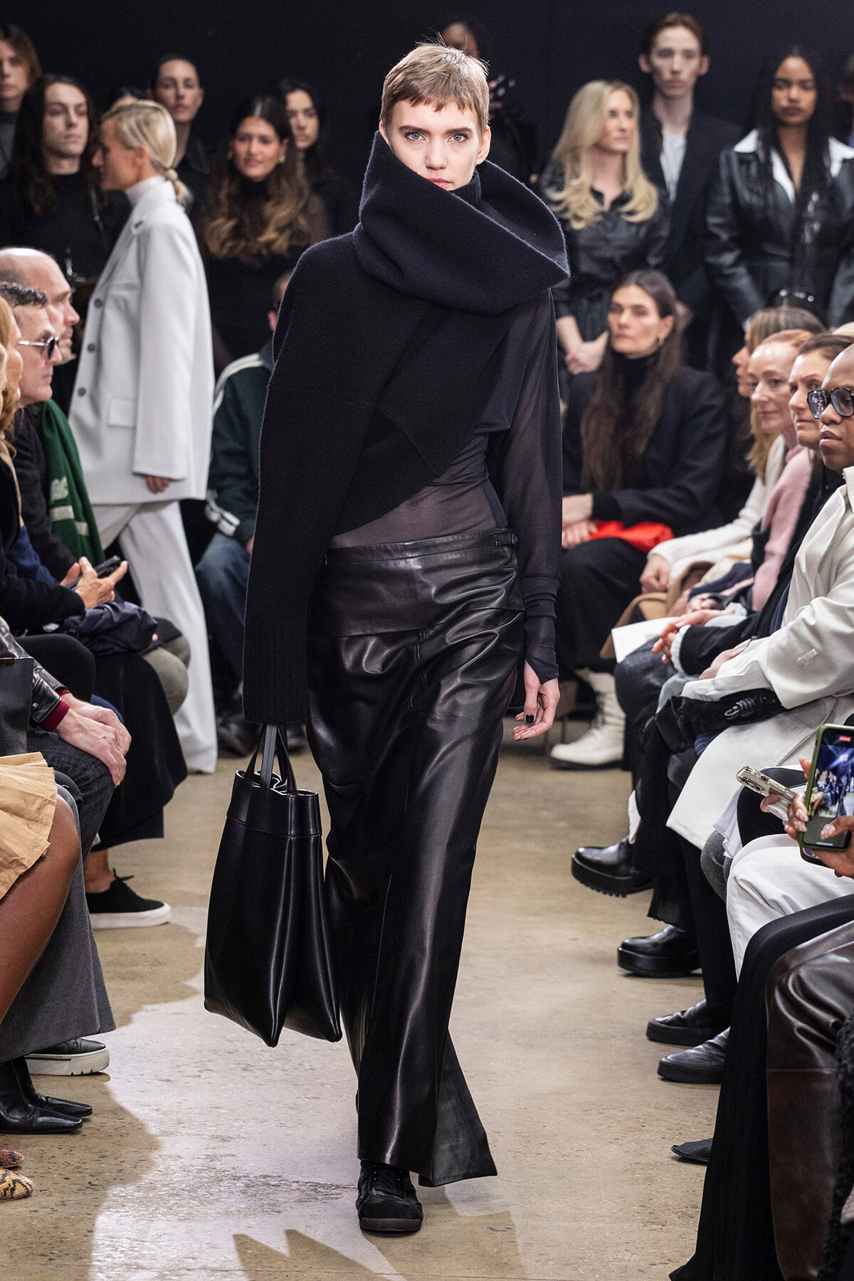 Model walks the runway in an all-black outfit with an oversized scarf, leather pants, and a black tote as the audience watches.