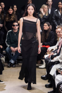 Model walks the runway in a black, layered dress as seated audience watches on both sides.