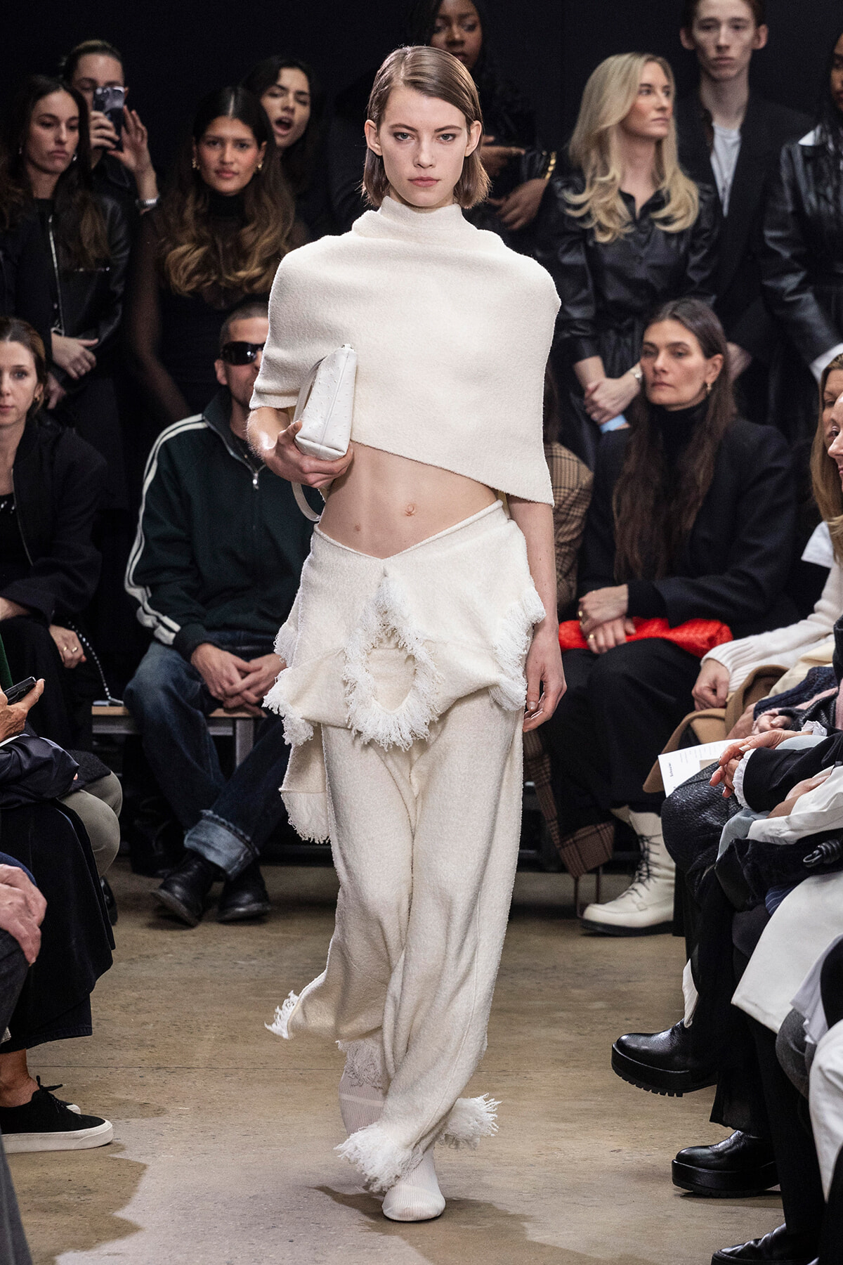 Model walks the runway in a white textured cape and layered frayed skirt, carrying a white clutch as a crowd watches.