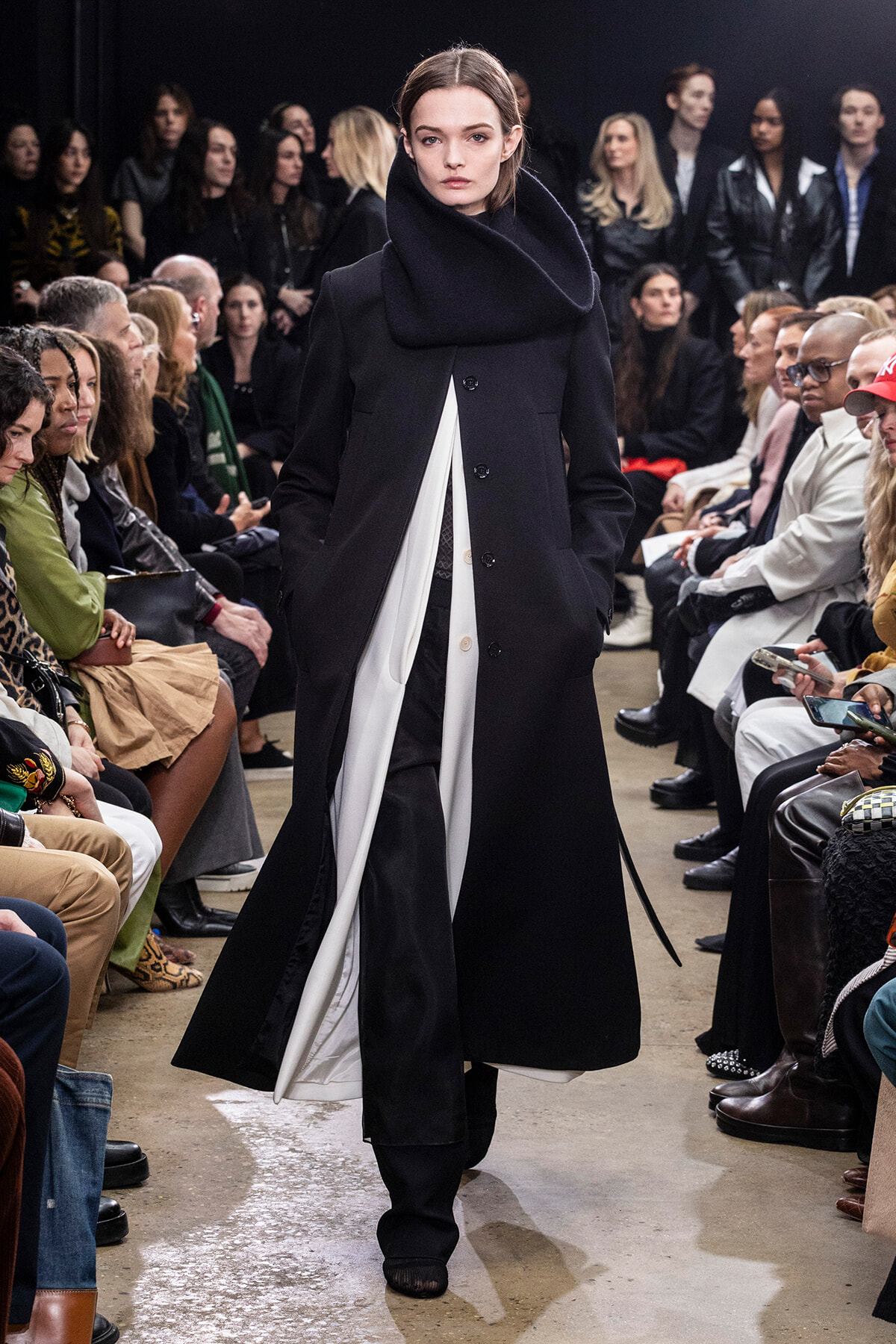Model walks the runway in an oversized black coat with a large scarf-like collar, surrounded by seated spectators.
