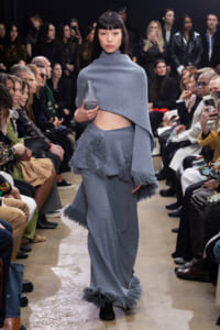 Model walks a fashion runway in a gray-blue textured knit cape and matching skirt, holding a small gray clutch as fringe details sway. Audience watches from both sides.