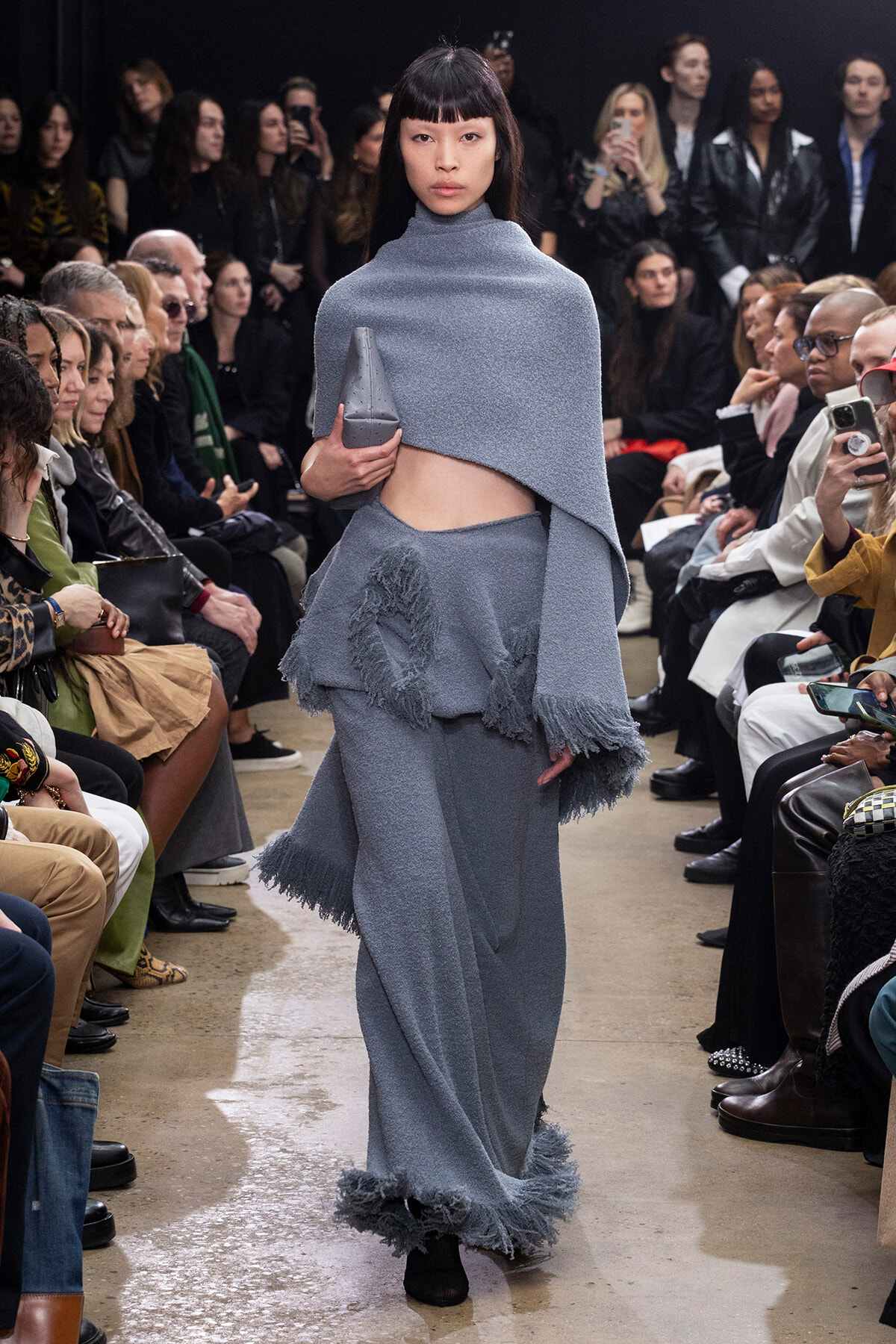 Model walks a fashion runway in a gray-blue textured knit cape and matching skirt, holding a small gray clutch as fringe details sway. Audience watches from both sides.