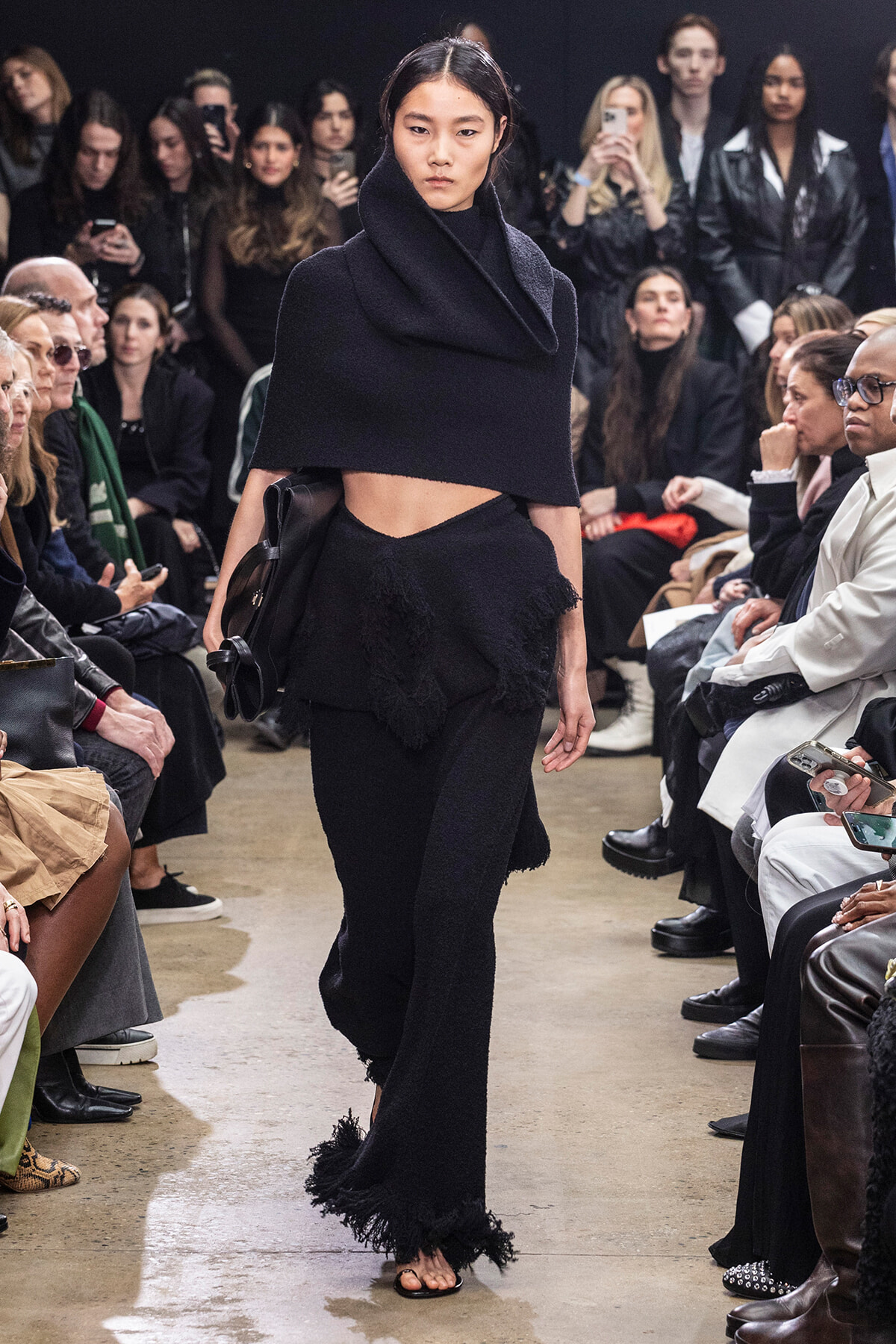 Model walks a fashion show wearing a black oversized cape top with a high collar and fringed black pants; audience watches from both sides.