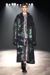Model walks the runway in a long black coat with green leaf patterns over a metallic silver dress, paired with black ankle boots.