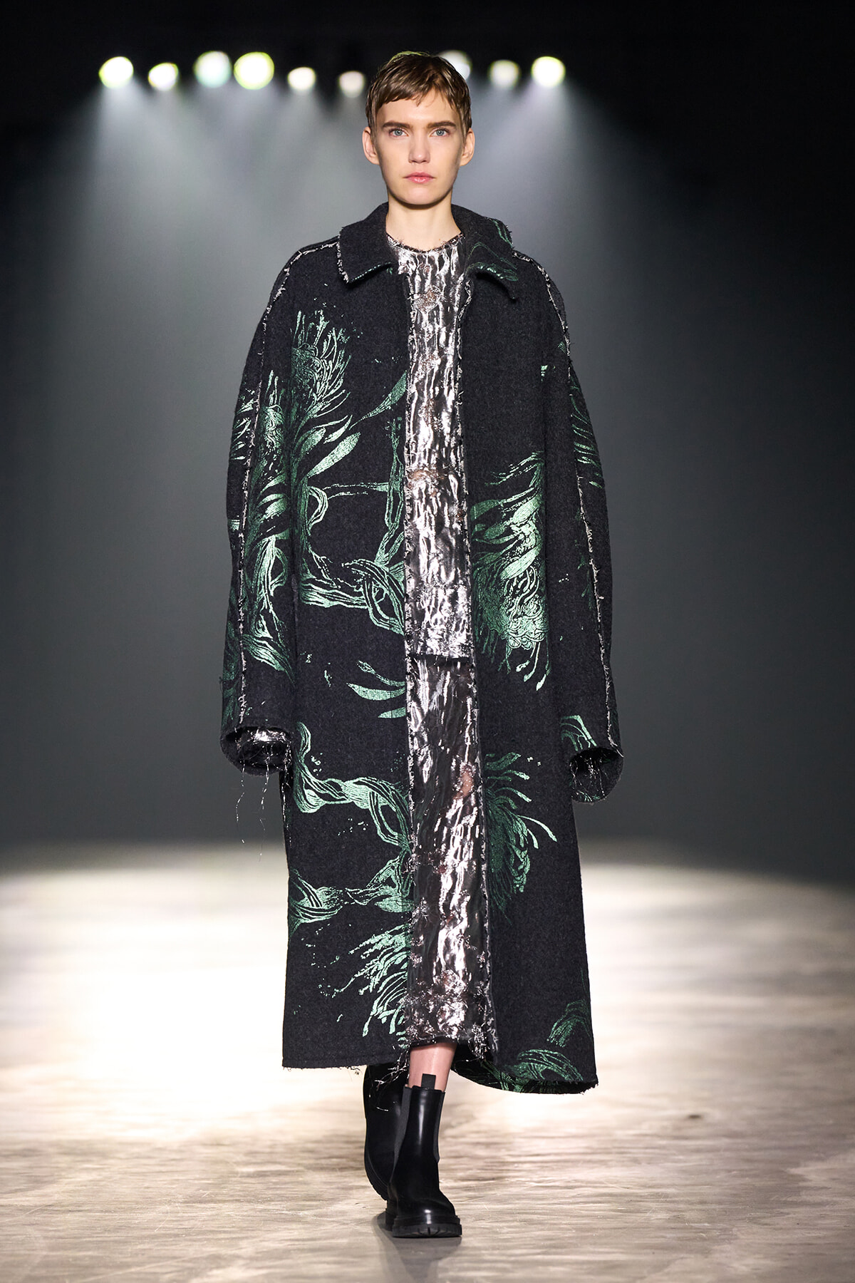 Model walks the runway in a long black coat with green leaf patterns over a metallic silver dress, paired with black ankle boots.