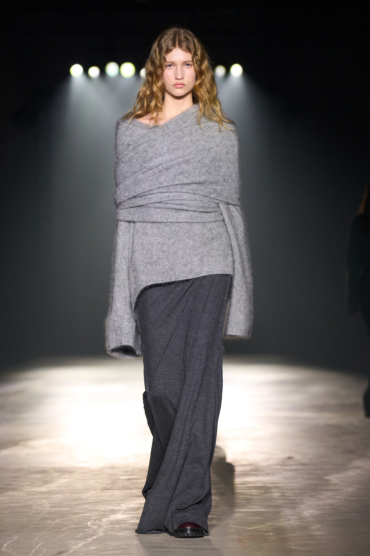 Model walks a fashion runway in a fuzzy light gray wrap sweater and wide-leg gray pants, spotlight behind casting a dramatic glow on the catwalk.