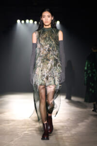 Model walking the runway in a sheer green floral dress, paired with gray elbow-length arm warmers and burgundy ankle boots.