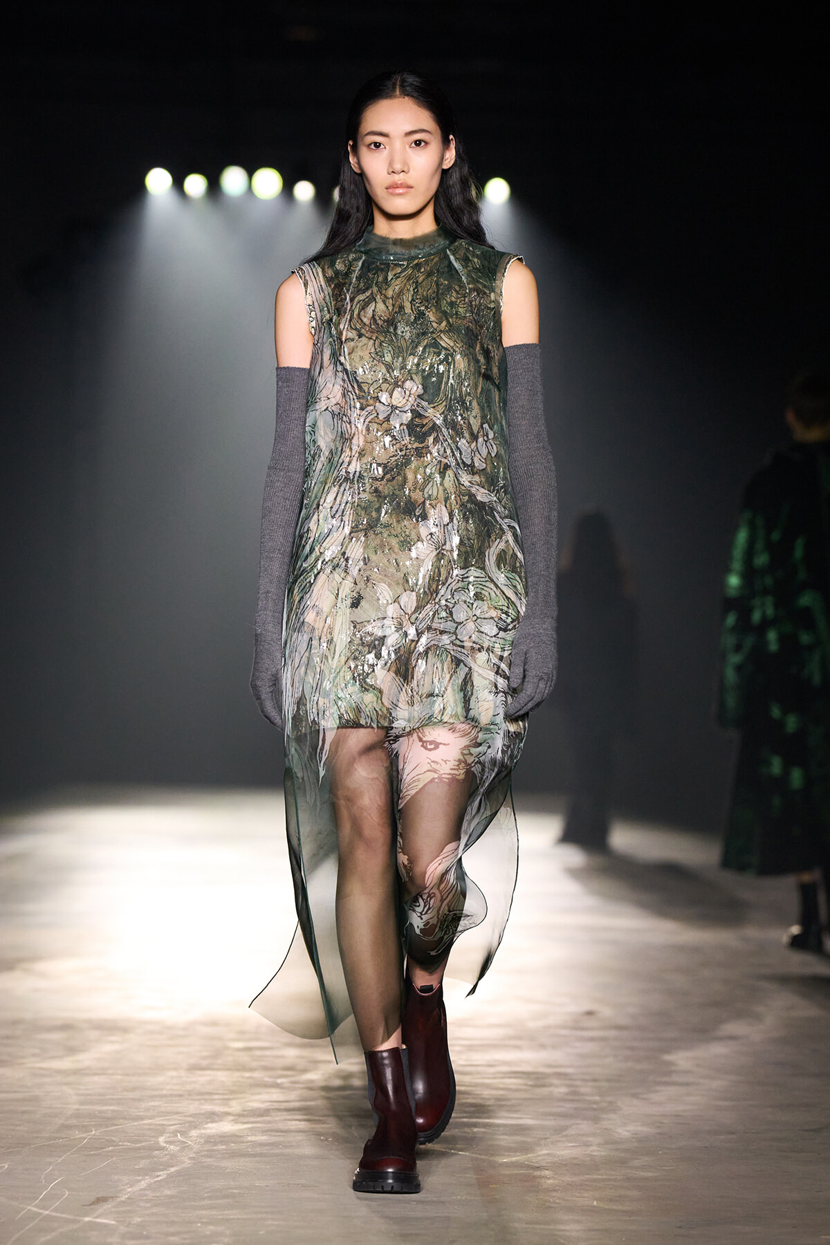Model walking the runway in a sheer green floral dress, paired with gray elbow-length arm warmers and burgundy ankle boots.