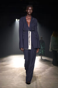Model walks the runway in a loose navy blazer and matching trousers with a white belt-like strip at the waist.