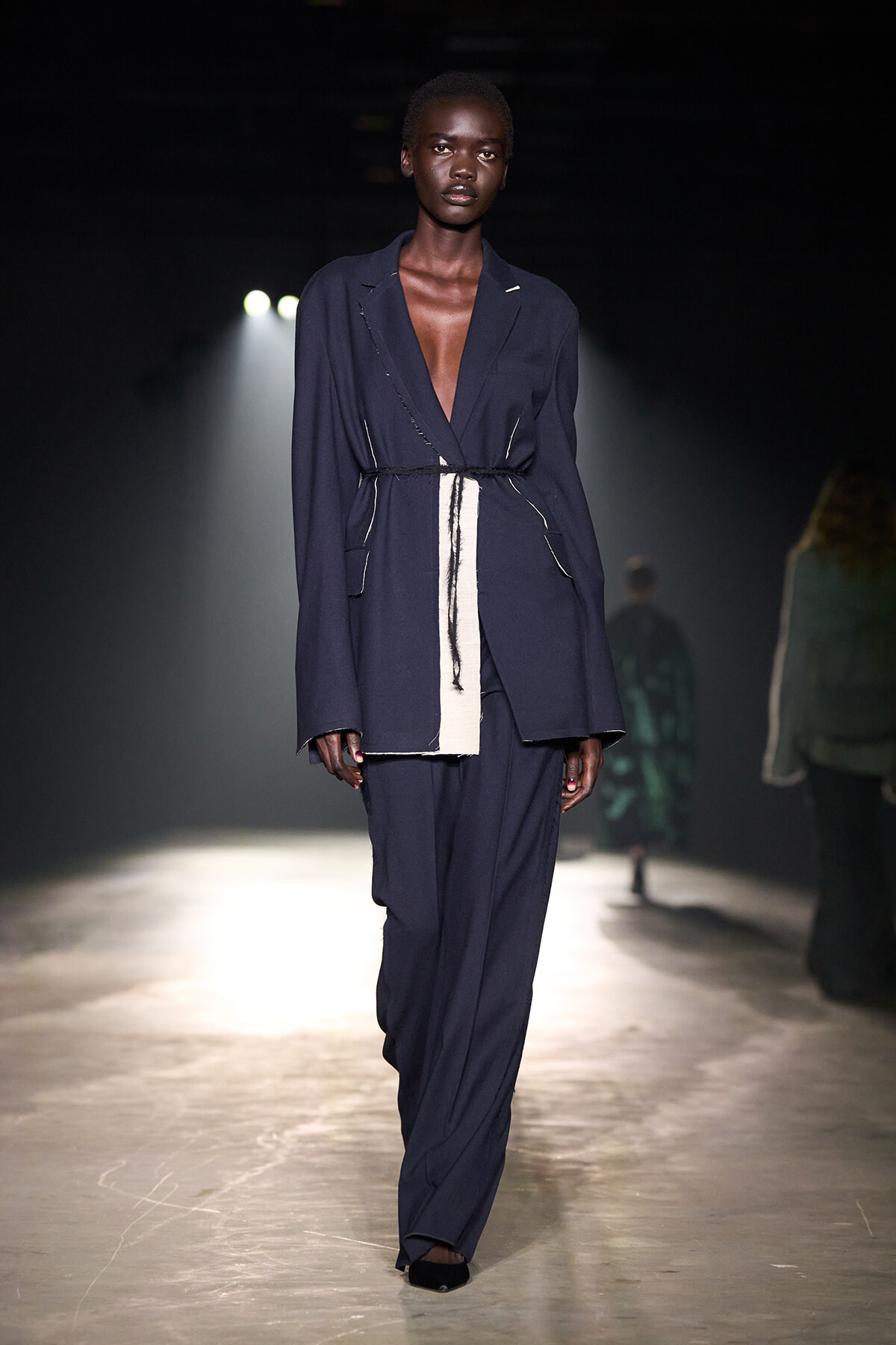 Model walks the runway in a loose navy blazer and matching trousers with a white belt-like strip at the waist.