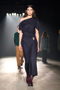 Model walks the runway in a navy off-shoulder jumpsuit with a small gold brooch at the hip; burgundy ankle boots add contrast.