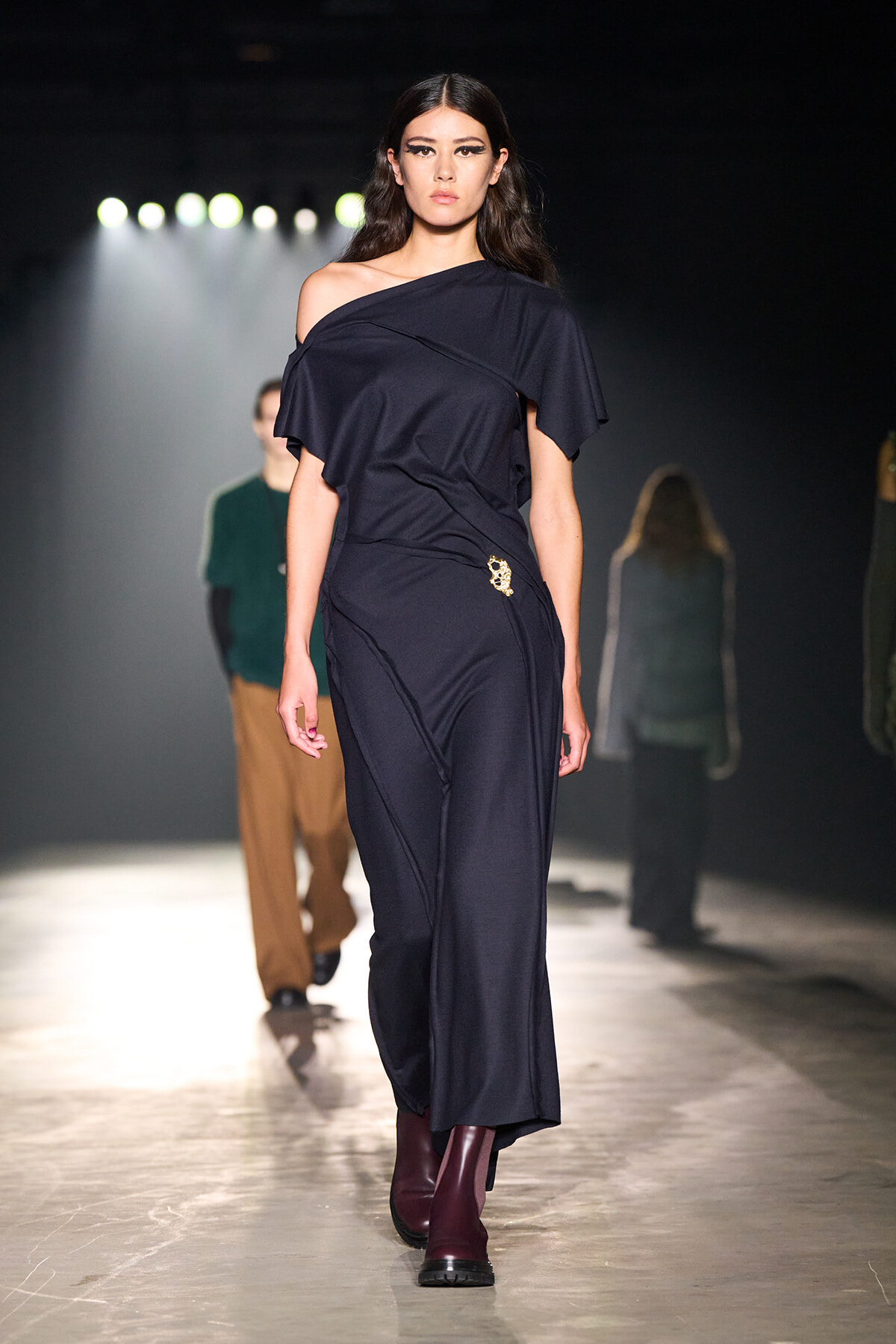 Model walks the runway in a navy off-shoulder jumpsuit with a small gold brooch at the hip; burgundy ankle boots add contrast.