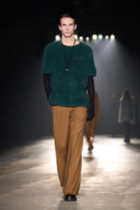 Male fashion model walking a runway in a dark venue, wearing a green fuzzy knit sweater, tan wide-leg pants, black gloves, and a pendant necklace.