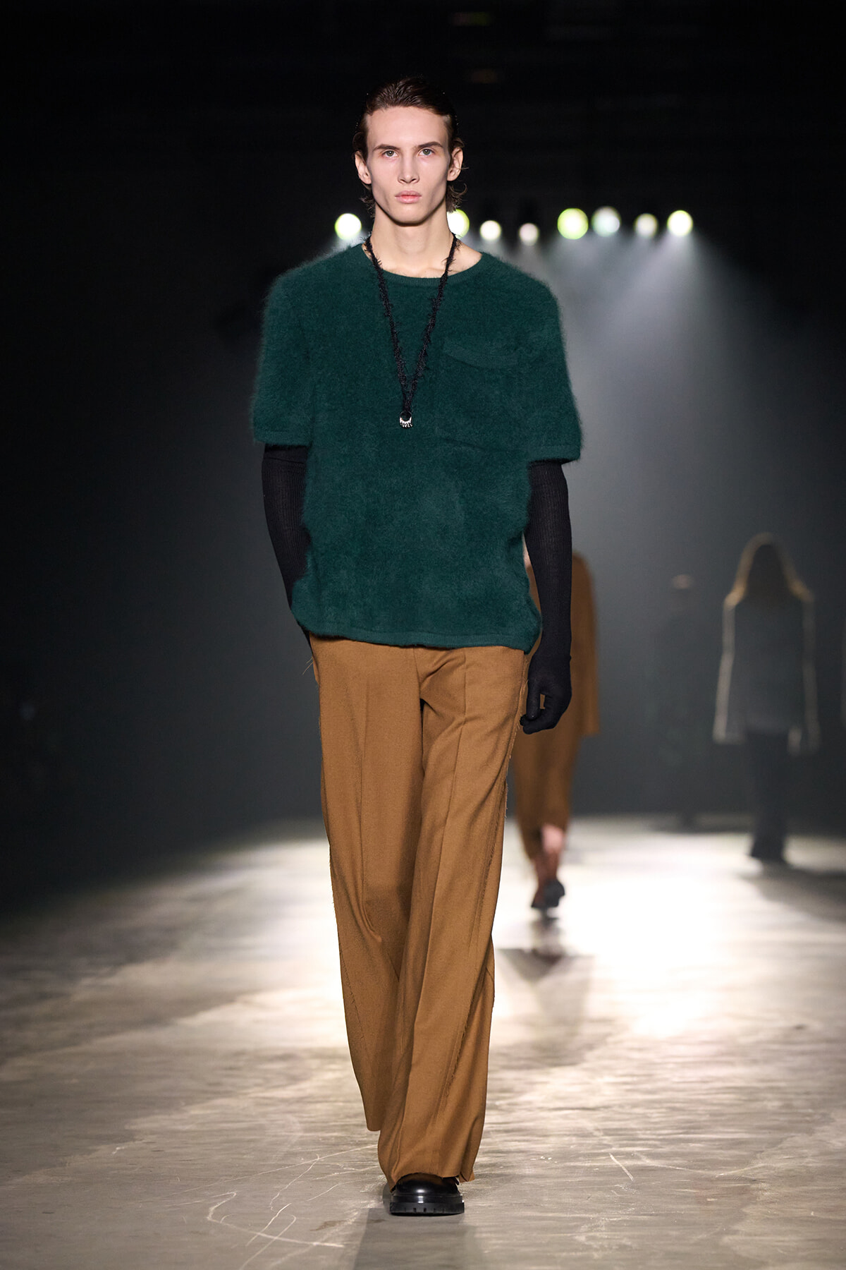 Male fashion model walking a runway in a dark venue, wearing a green fuzzy knit sweater, tan wide-leg pants, black gloves, and a pendant necklace.