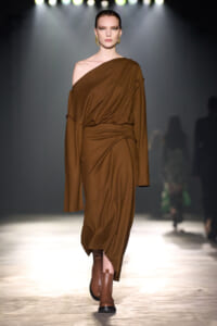 Model walking runway in a brown one-shoulder draped dress and brown ankle boots under spotlight.