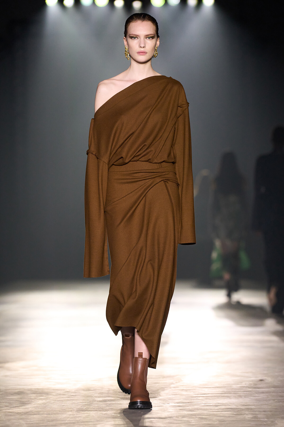 Model walking runway in a brown one-shoulder draped dress and brown ankle boots under spotlight.