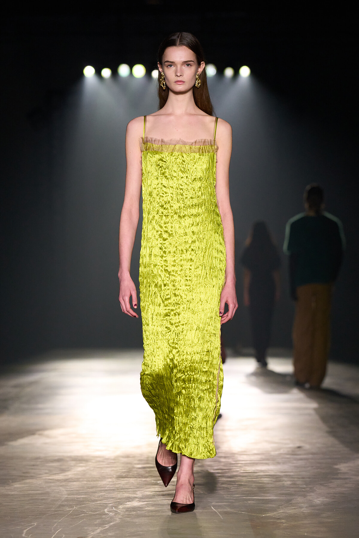 Model walks a runway in a bright lime-green, textured slip dress with thin straps and gold earrings under spotlight.