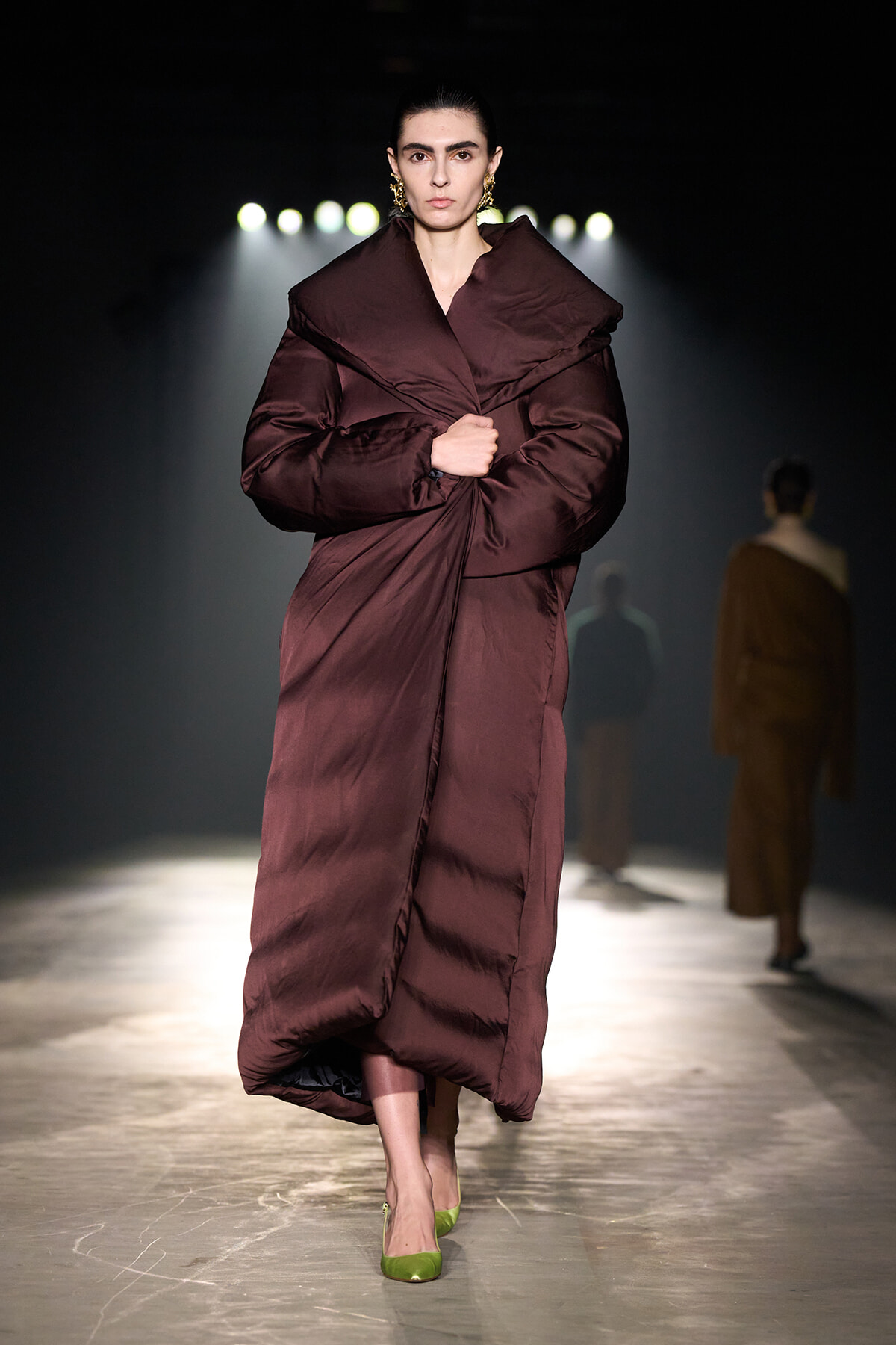 Model walking a runway in a oversized burgundy puffer coat, bright backstage lights in the background, wearing lime green shoes.