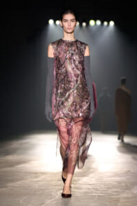 Model walks a fashion runway wearing a sheer pink floral dress with off-the-shoulder cutouts and long gray knit gloves, under bright backstage spotlights.