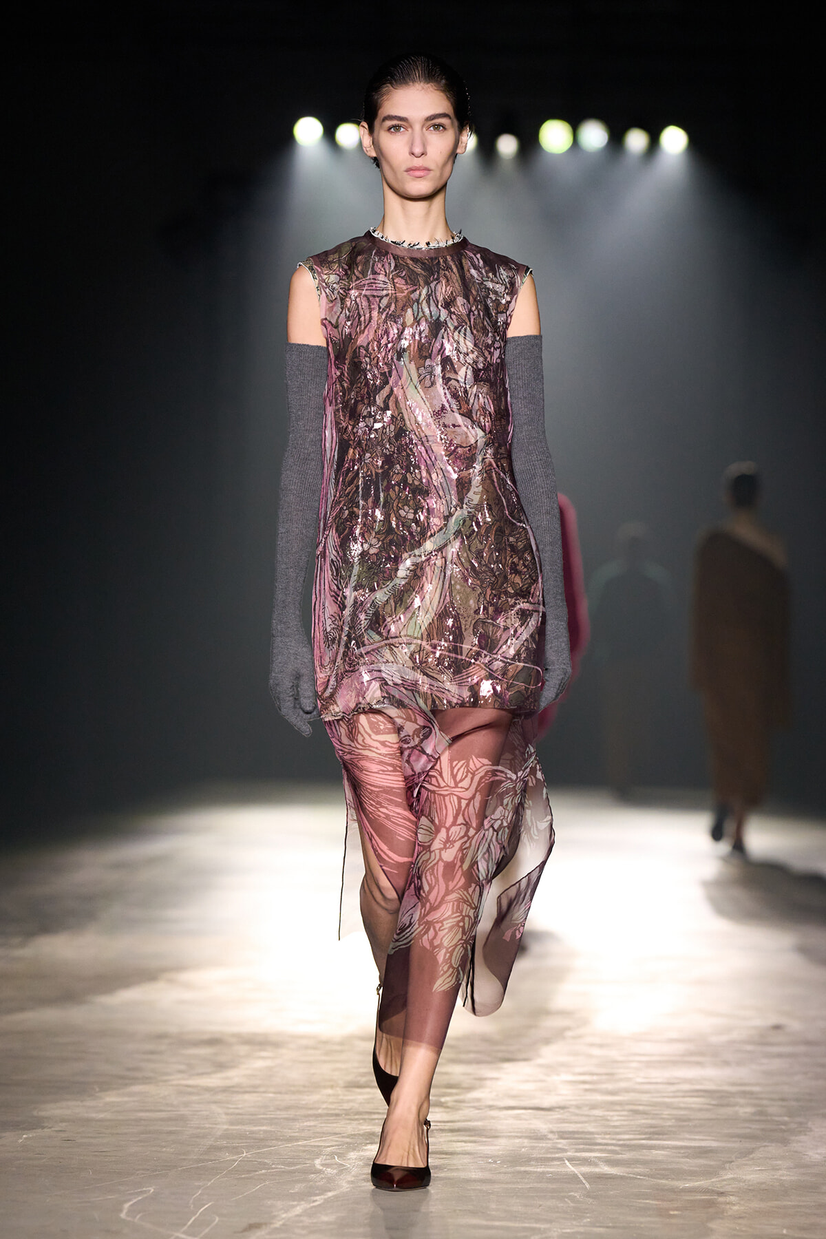 Model walks a fashion runway wearing a sheer pink floral dress with off-the-shoulder cutouts and long gray knit gloves, under bright backstage spotlights.
