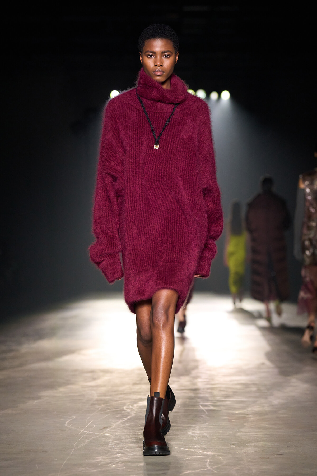 Model walks the runway in an oversized burgundy fuzzy knit turtleneck sweater with a pendant necklace.