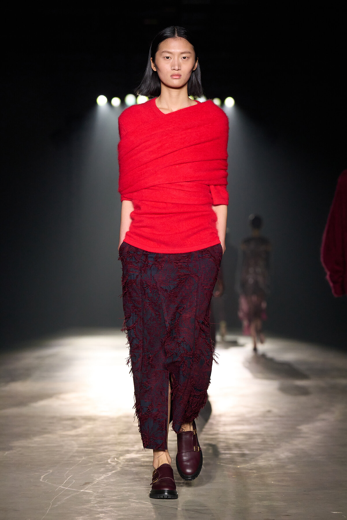 Runway model walks toward the camera in a bright red wrapped top and burgundy lace skirt.