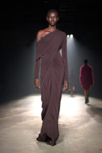 Model walks a runway wearing a one-shoulder mauve draped gown, spotlight behind her in a dark venue.