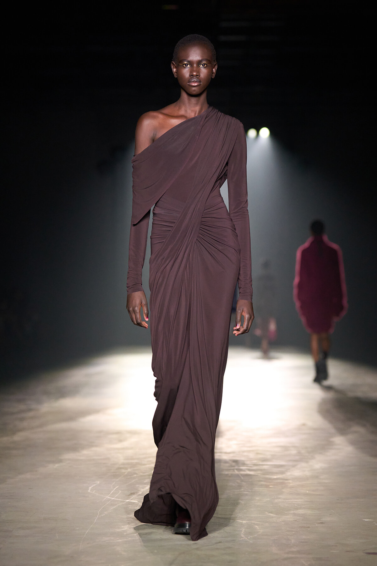 Model walks a runway wearing a one-shoulder mauve draped gown, spotlight behind her in a dark venue.