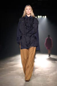 Female model walking a runway in a navy blouse with black embroidery and camel wide-leg pants under spotlight.