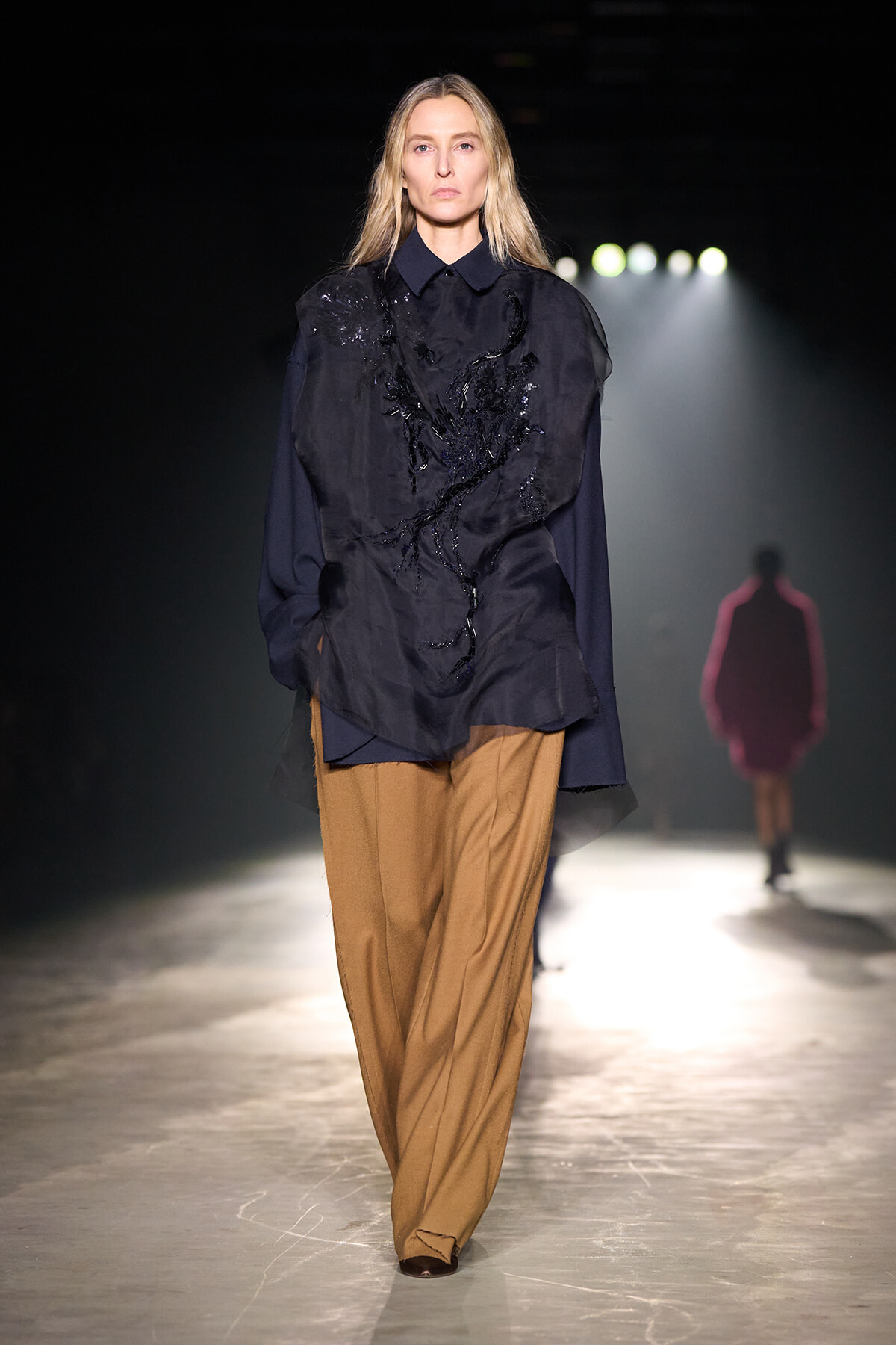 Female model walking a runway in a navy blouse with black embroidery and camel wide-leg pants under spotlight.