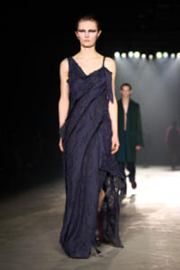 Model walks the runway in a navy blue, frayed-ragged dress under bright spotlights.
