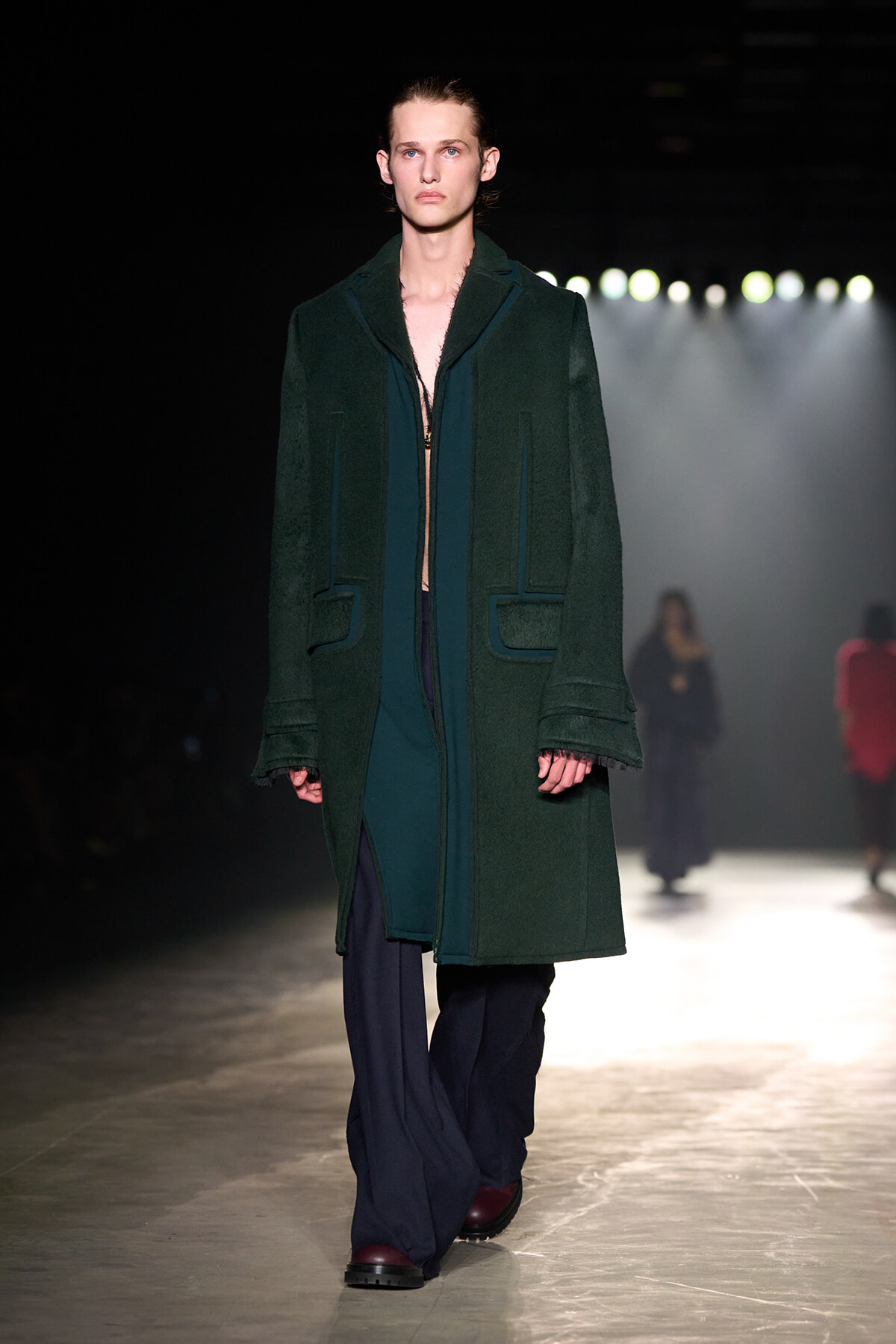 Model walking a runway in a long dark green coat over a teal outfit, on a dimly lit catwalk with spotlights in the background