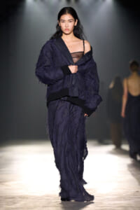 Model walking a fashion runway in a dark navy textured suit with a sheer black top visible at the neckline and an oversized jacket.