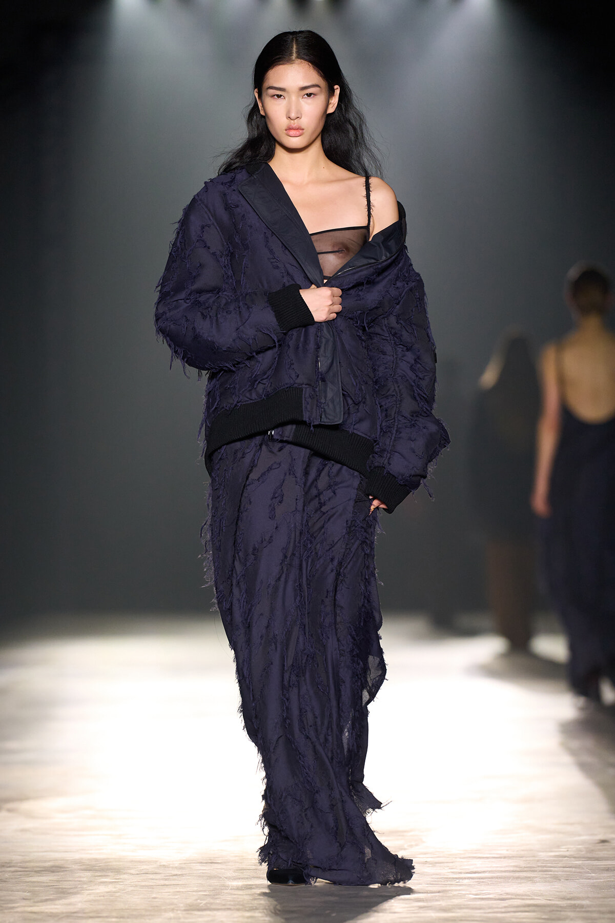Model walking a fashion runway in a dark navy textured suit with a sheer black top visible at the neckline and an oversized jacket.
