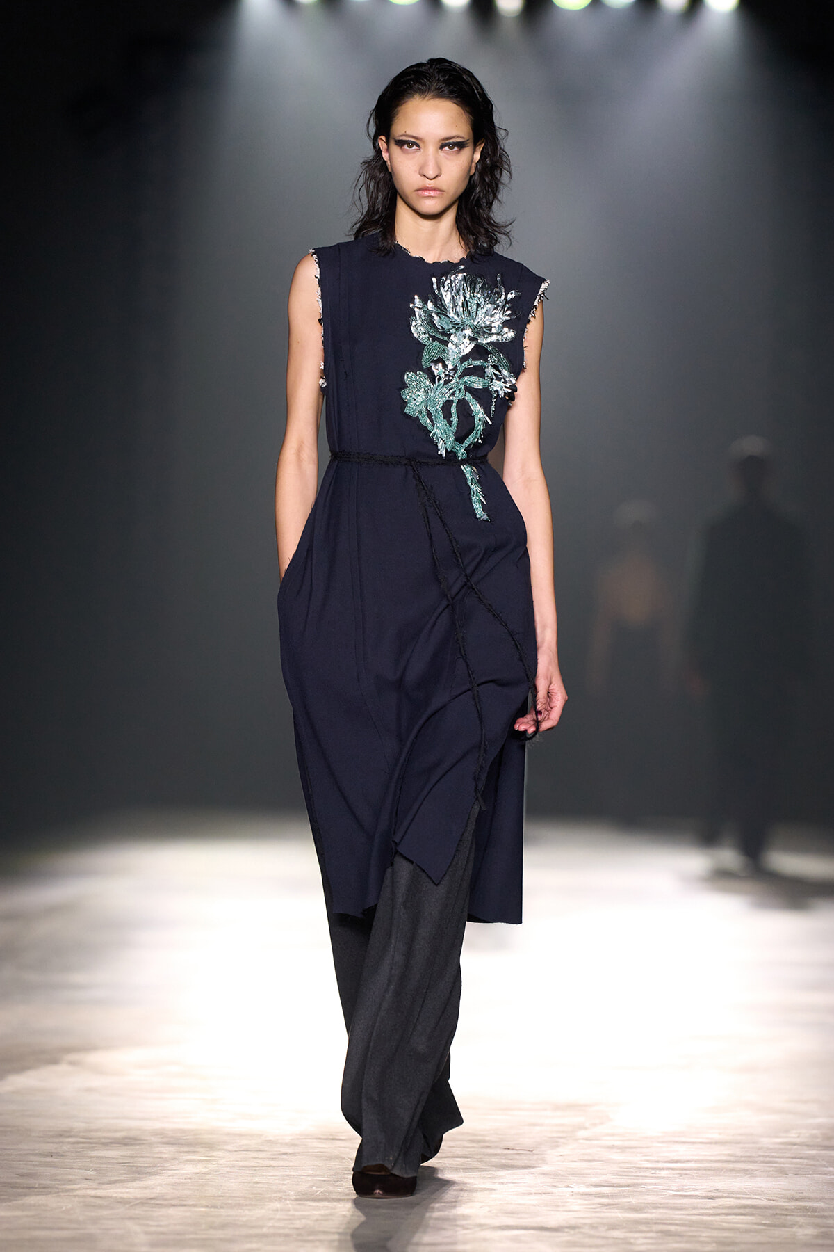Model walking a fashion runway in a sleeveless navy dress with teal floral embroidery, a cinched waist, and layered detailing under bright spotlights.