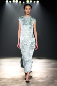 Model walking the runway in a pale blue textured velvet dress with cap sleeves under bright stage lights