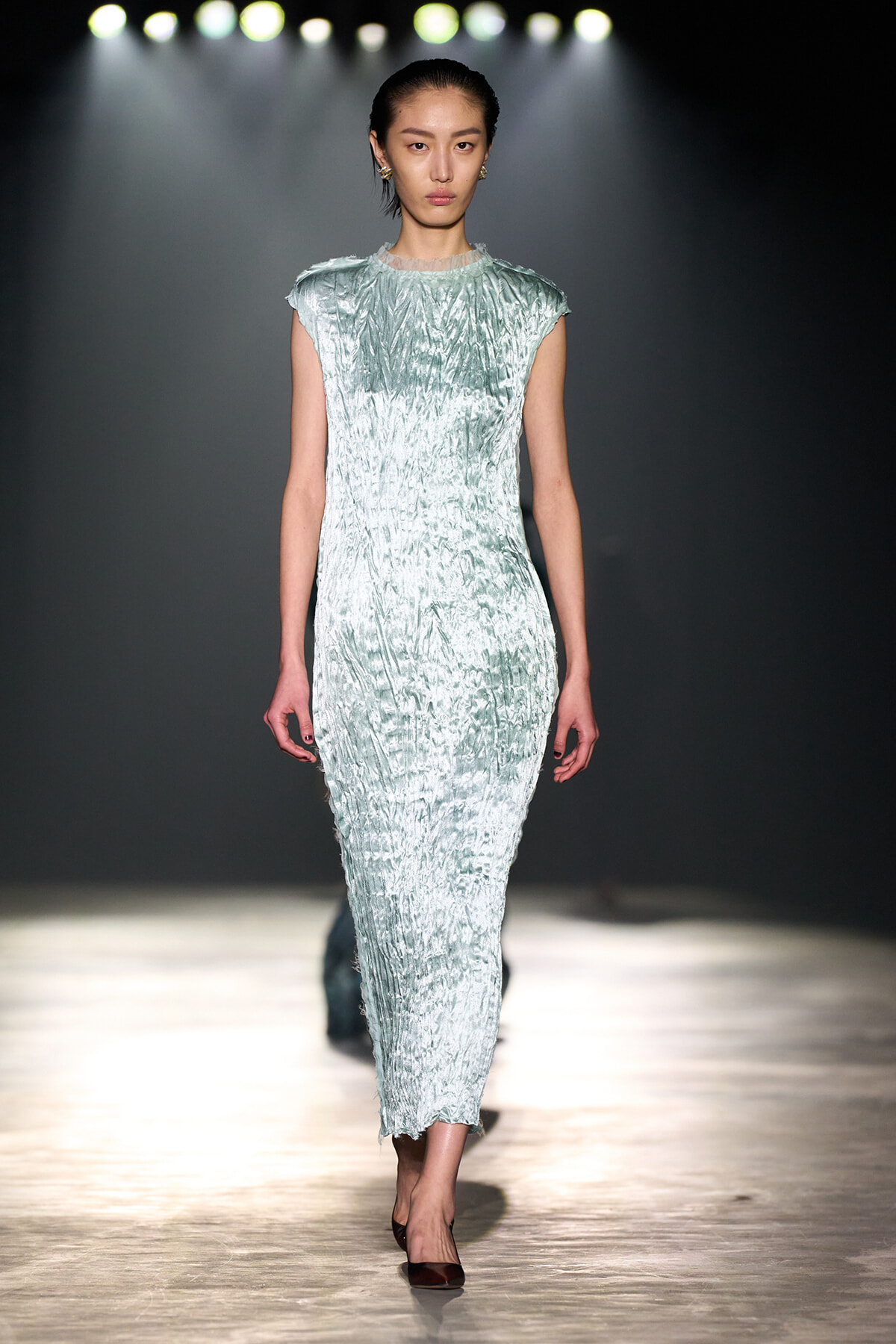 Model walking the runway in a pale blue textured velvet dress with cap sleeves under bright stage lights