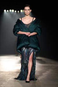 Model walking a runway in a deep emerald oversized quilted wrap over a sheer, patterned gown; arms crossed, bright spotlights behind her.