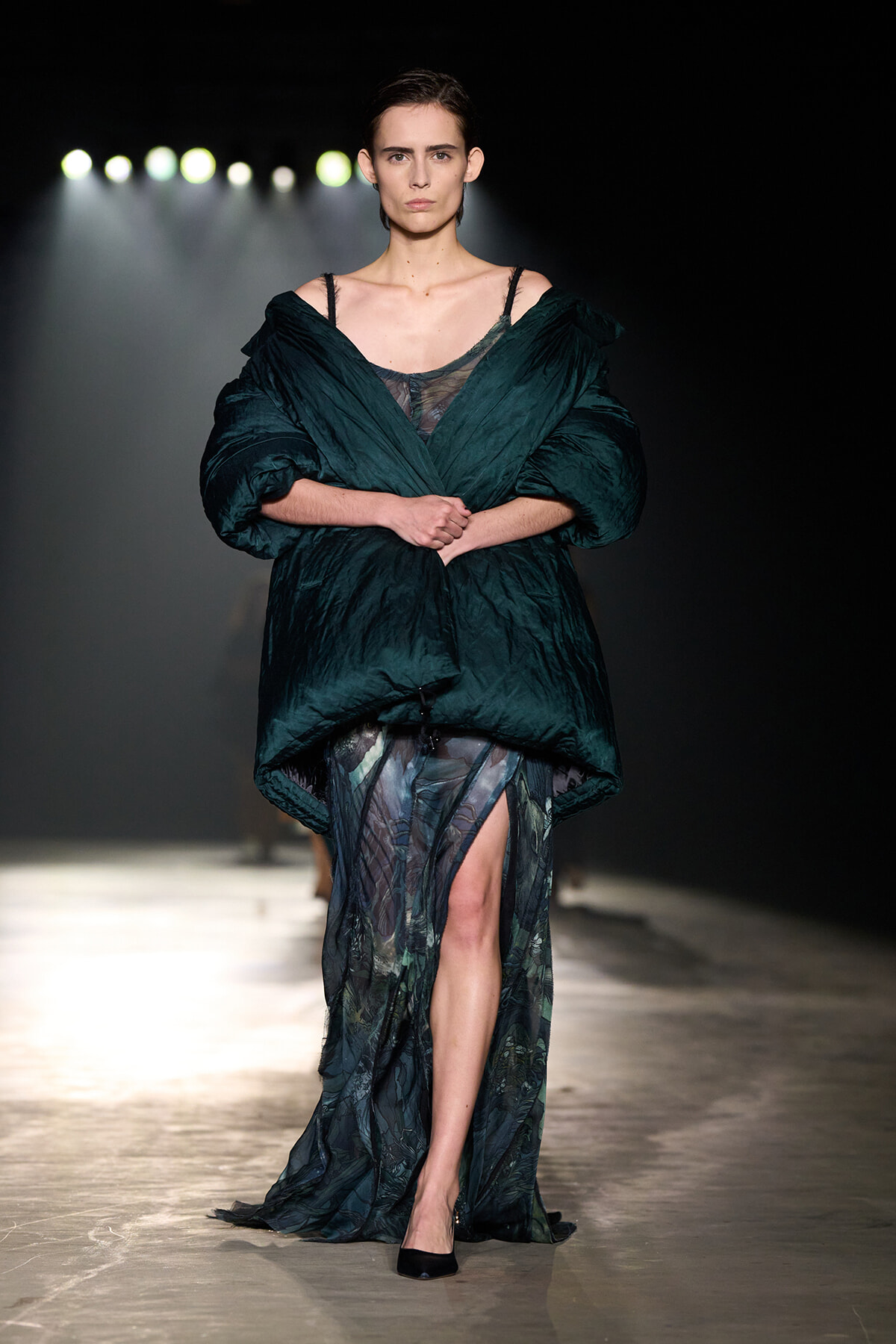 Model walking a runway in a deep emerald oversized quilted wrap over a sheer, patterned gown; arms crossed, bright spotlights behind her.