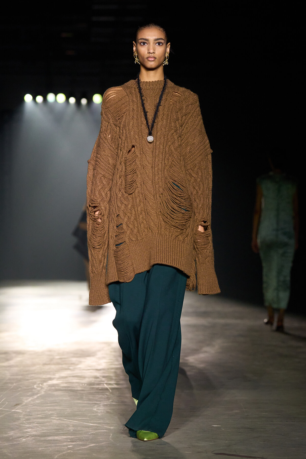 Runway model wearing an oversized brown cable-knit sweater with distressed holes, teal wide-leg pants, and gold jewelry; spotlighted catwalk scene.