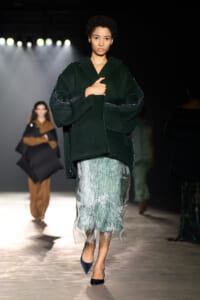 Model on a runway wearing an oversized dark green coat with seam detailing and a mint-green patterned skirt, walking toward the camera with blue heels.