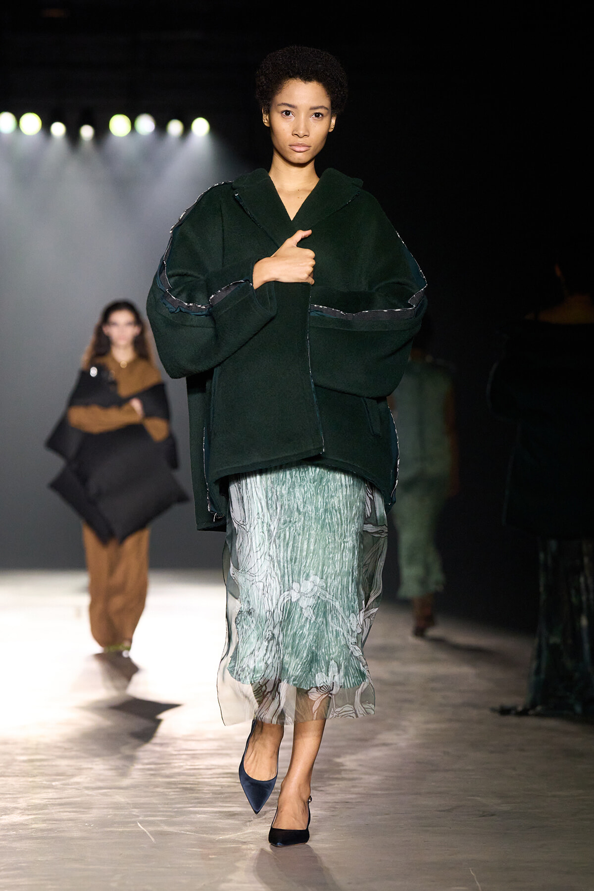 Model on a runway wearing an oversized dark green coat with seam detailing and a mint-green patterned skirt, walking toward the camera with blue heels.