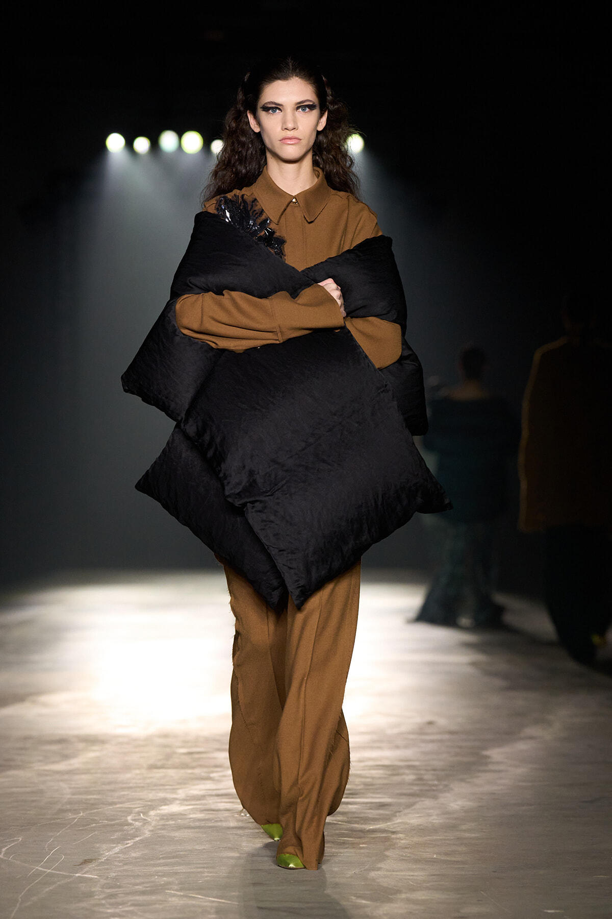 Model walking a runway in a brown suit with an oversized black quilted wrap, wearing lime-green shoes under bright stage lights.