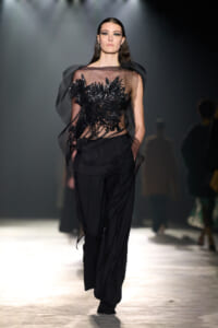 Model on a runway wearing a sheer black top with black sequin embroidery and black tailored pants.