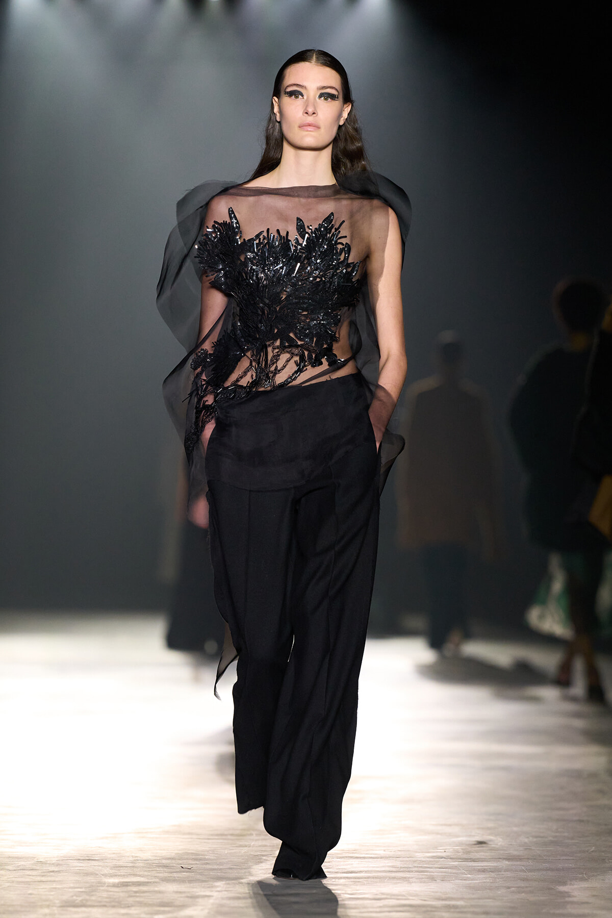 Model on a runway wearing a sheer black top with black sequin embroidery and black tailored pants.