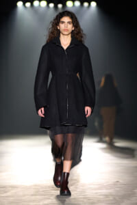 Model walking down a fashion show runway in a black belted coat and dark sheer skirt under bright runway lights.