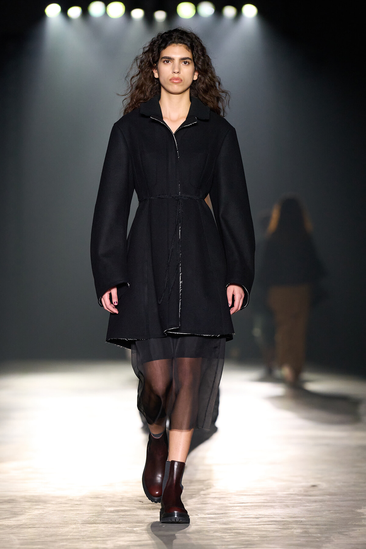 Model walking down a fashion show runway in a black belted coat and dark sheer skirt under bright runway lights.