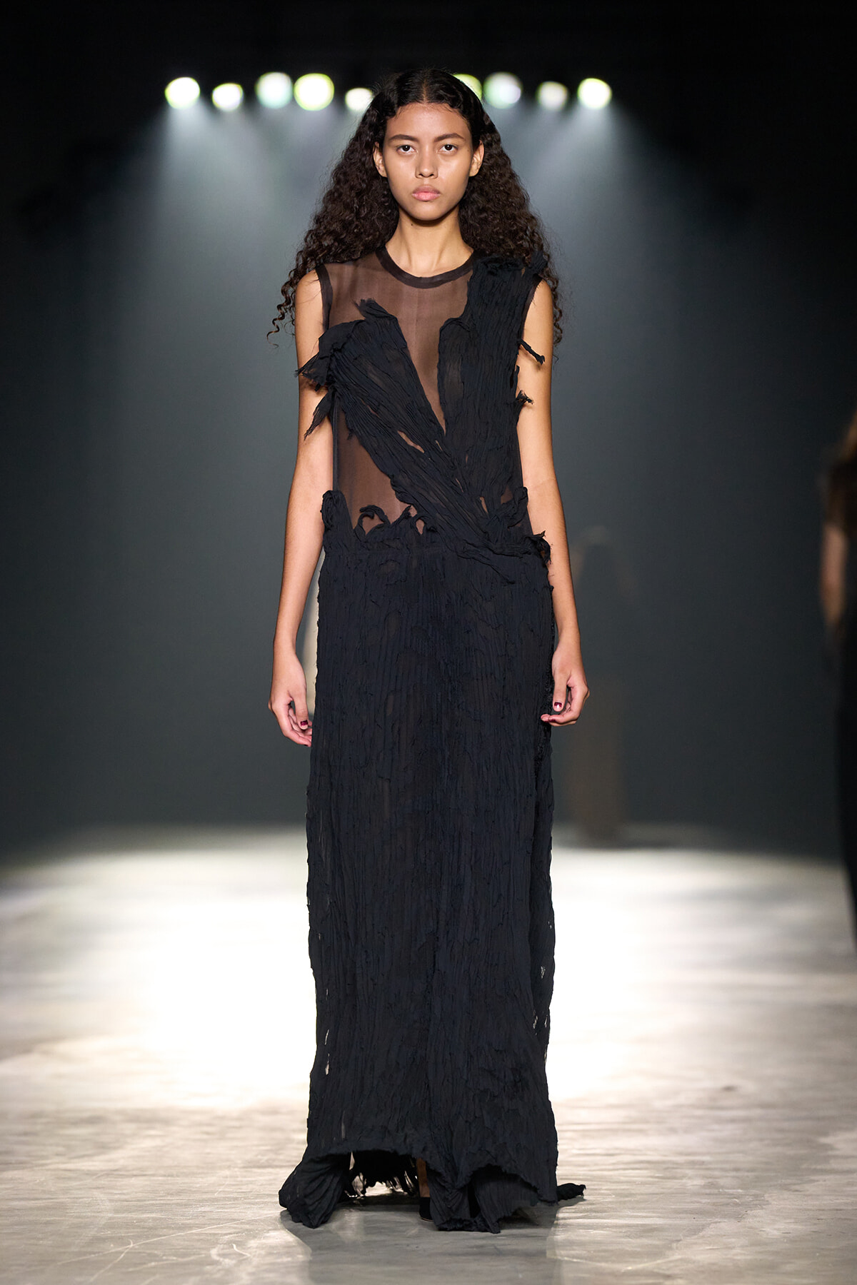 Model on a fashion runway wearing a long black sheer gown with shredded fringe details under bright stage lights.